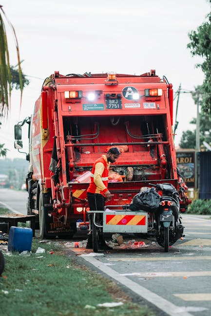 A waste collection worker wearing an orange and yellow uniform is operating a red rubbish truck, which is parked on the roadside. The truck's rear hopper is open, revealing a metal calibration plate a