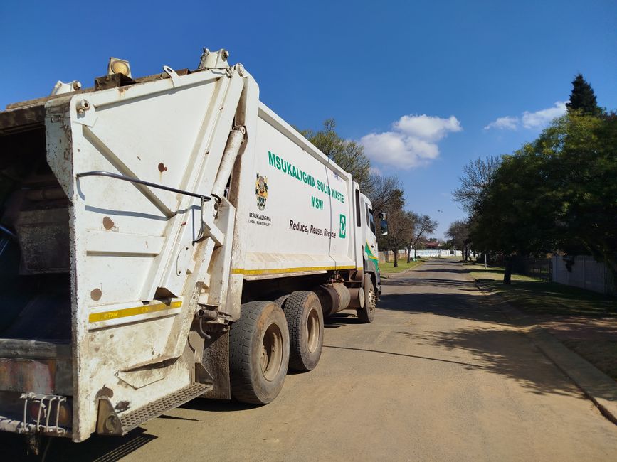A waste collection worker wearing an orange and yellow uniform is operating a red rubbish truck, which is parked on the roadside. The truck's rear hopper is open, revealing a metal calibration plate a